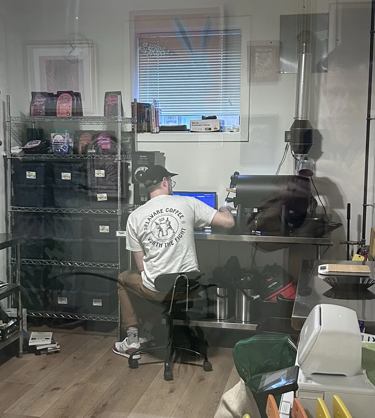Adam Rausch sits at a small drum roaster in a back room, in a T-shirt that reads 'Delaware Coffee, Roasters of Exotic Origins, Roasted in Wilmington, Worth the Fight, 2023,' with shelves of bagged coffee behind him.