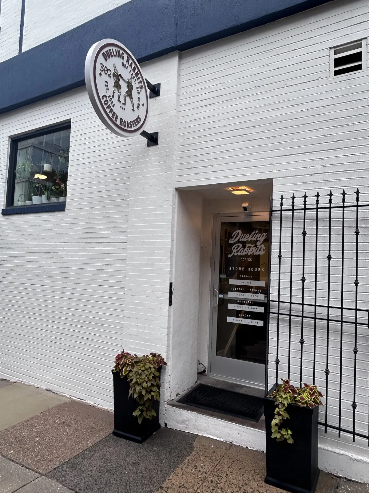 A round white sign reading 'Dueling Rabbits 302 Coffee Roasters, Hop Fast Sip Slow, Est 2023,' with two boxing rabbits in brown gloves, hung above a white brick entrance.